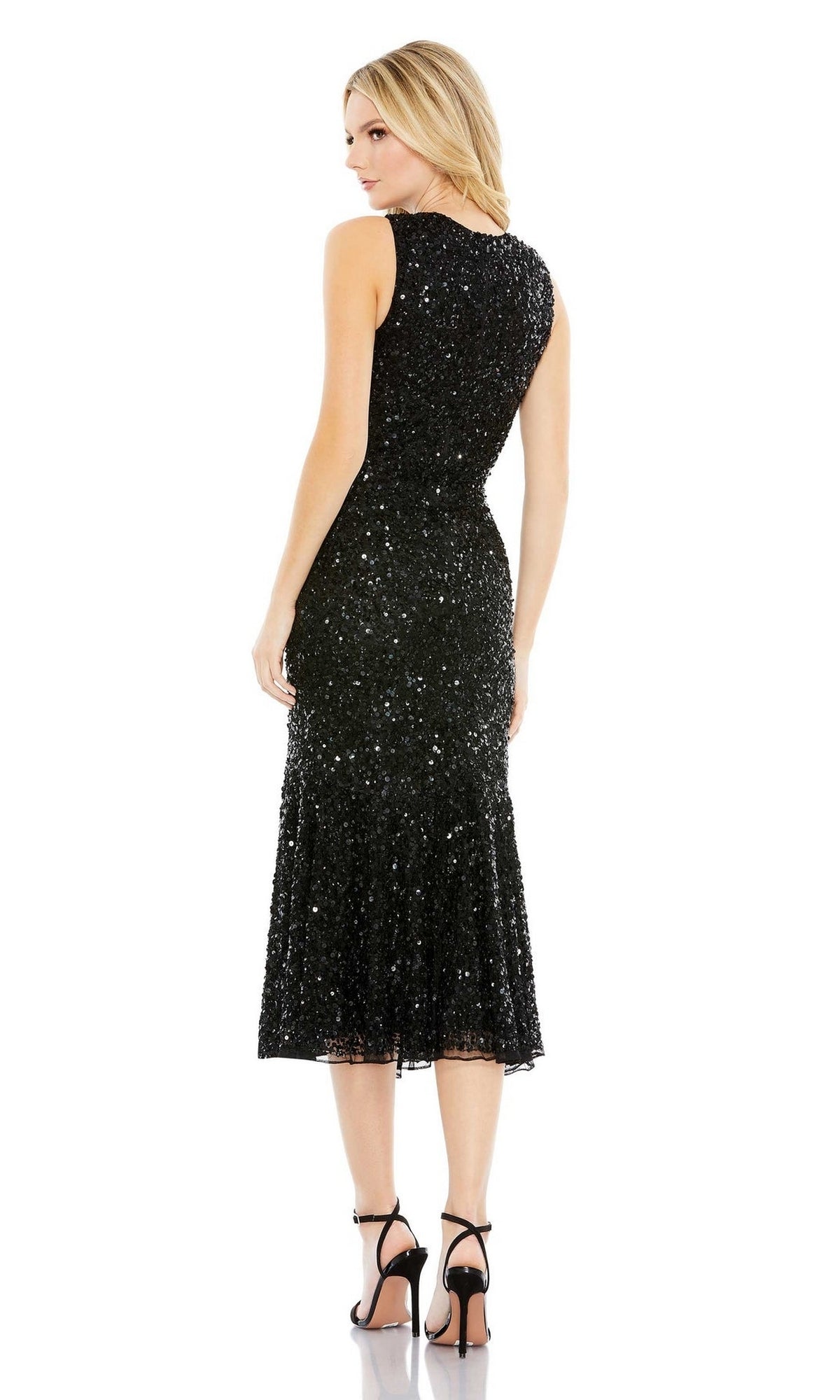 A woman stands facing away, wearing the Short Semi-Formal Dress 5597 by Mac Duggal—a sleeveless, high-neck sequin dress with a flared hem—paired with black high-heeled sandals.